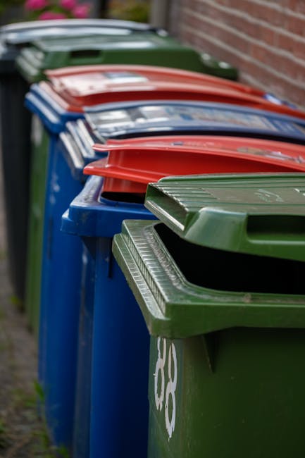 A row of large, wheelie-style bins with contrasting colours of green, blue, red, and dark green are lined up along a pavement beside a brick wall. The bins are made of durable plastic with textured li