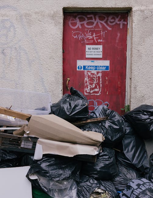 A pile of discarded rubbish and black garbage bags is stacked in front of a red metal door set into a rough beige wall. The rubbish includes a mixture of black plastic bags, cardboard, and other waste